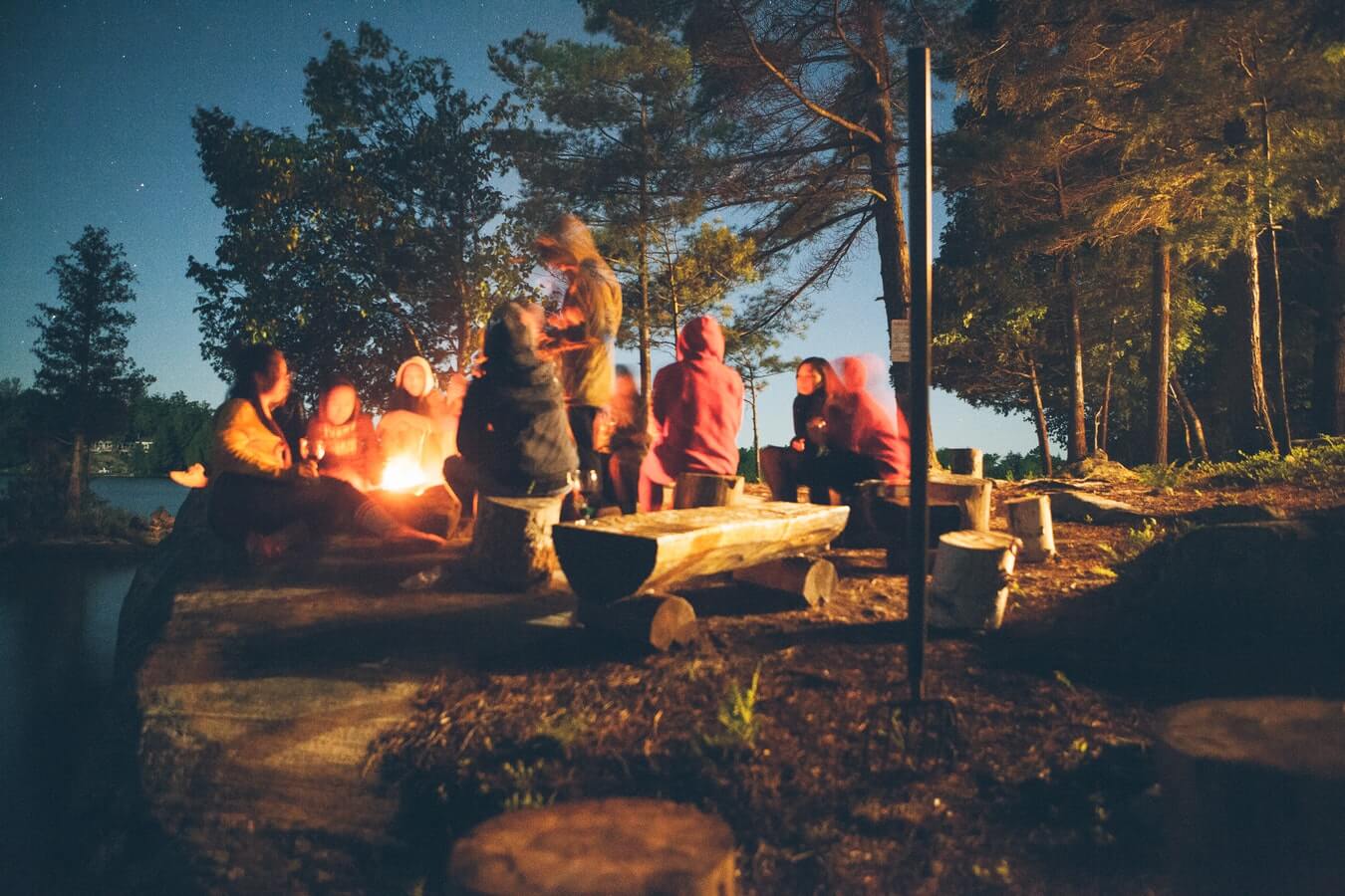 A group outside sitting around a bonfire on the edge of a lake in an early evening.