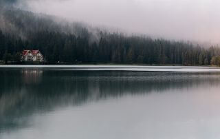 A beautiful cottage sits on the edge of a wooded hillside, seen from across a foggy lake on a misty day.