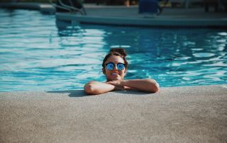 Smiling girl wearing sunglasses is inside a swimming pool leaning against the side.