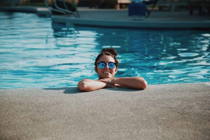 Smiling girl wearing sunglasses is inside a swimming pool leaning against the side.