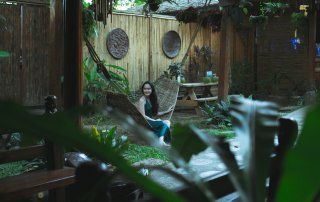 A woman sits in a hammock in a lush, green backyard with a stone patio.