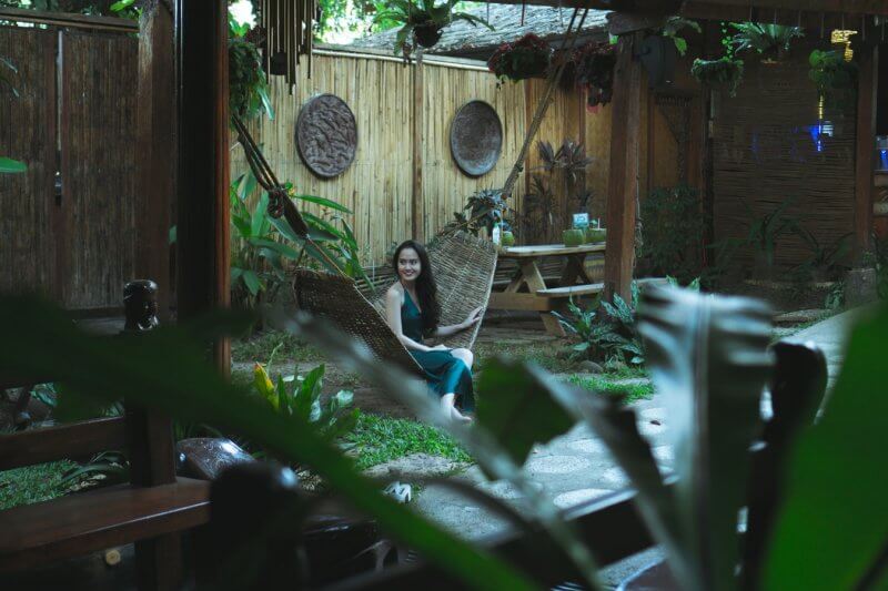 A woman sits in a hammock in a lush, green backyard with a stone patio.
