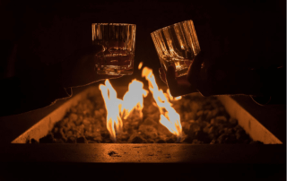 two people cheers their whiskey glasses over an outdoor fire pit