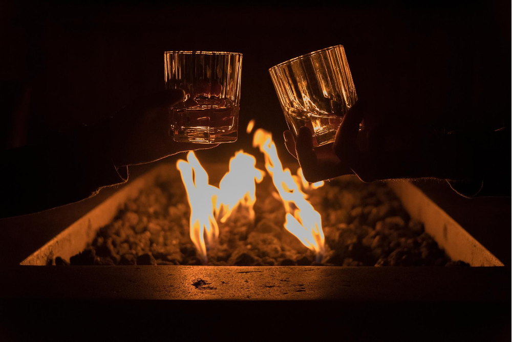 two people cheers their whiskey glasses over an outdoor fire pit